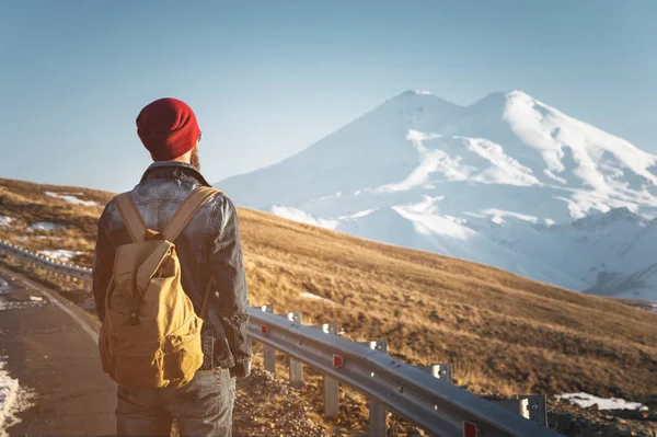 Bearded tourist hipster man in sunglasses with a backpack stand back on a roadside bump and watching the sunset against the background of a snow capped mountain — Stock Photo, Image