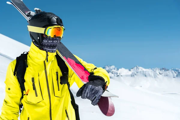 A portrait of a skier in a protective helmet and glasses is a mask and scarf with skis on his shoulder in the snow-capped mountains of the Caucasus. Skiing, — Stock Photo, Image