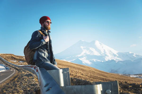 Skäggiga turist hipster man i solglasögon med en ryggsäck som sitter på en vägkanten bula och titta på solnedgången mot bakgrund av ett snöklädda berg — Stockfoto