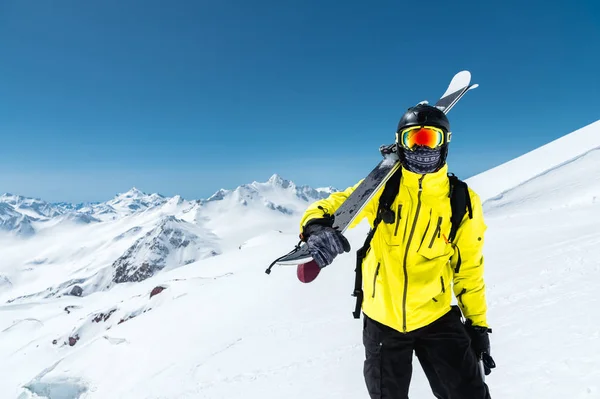 Um retrato de um esquiador em um capacete protetor e óculos é uma máscara e um lenço com esquis em seu ombro nas montanhas cobertas de neve do Cáucaso. Esqui , — Fotografia de Stock