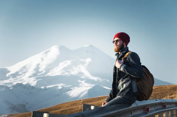 Bearded tourist hipster man in sunglasses with a backpack sitting on a roadside bump and watching the sunset against the background of a snow-capped mountain — Stock Photo, Image