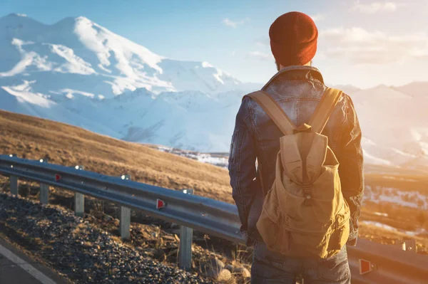 Barbuto turista hipster uomo in occhiali da sole con uno zaino stare indietro su un dosso della strada e guardare il tramonto sullo sfondo di una montagna innevata — Foto Stock