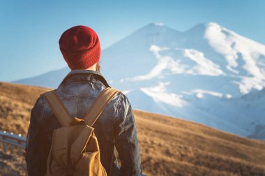 Sakallı turist hipster erkek güneş gözlüğü bir sırt çantası ile dimdik geri üzerinde yol kenarına yerleştirilen bir yumru ve şapkalı kar dağ çerçevede gün batımını izlemek