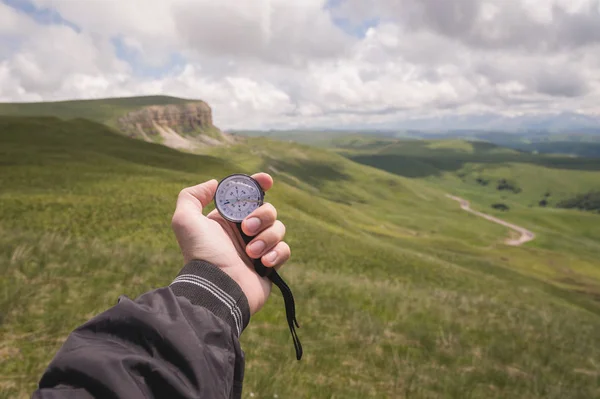 A mans hand of a tourist with an authentic compass on the background of a mountain road landscape — Stock Photo, Image