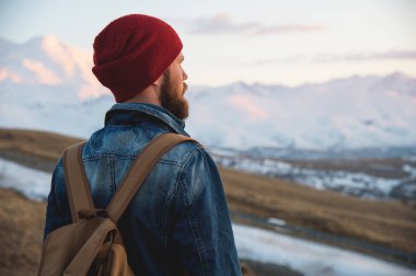 Sakallı turist hipster adam şapka ile bir sırt çantası içinde durmak geri yol kenarına yerleştirilen bir yumru ve şapkalı kar dağ çerçevede gün batımını izlemek