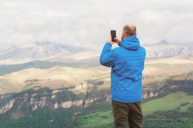 Sakallı bir adam bir membran mavi ceketli ve dağların fon karşı doğada standları telefon manzara fotoğraf çeker