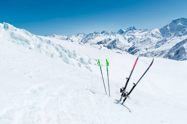 Um par de esquis e bastões de esqui sobressaem na neve na encosta da montanha do Cáucaso contra o pano de fundo da cordilheira caucasiana e o céu azul em um dia ensolarado — Fotografia de Stock