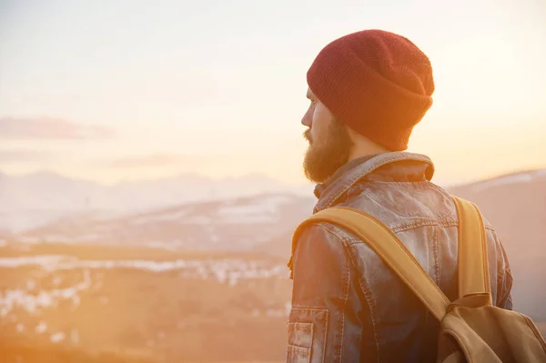 Barbuto turista hipster uomo in un cappello con uno zaino stand back su un dosso della strada e guardando il tramonto sullo sfondo di una montagna innevata — Foto Stock