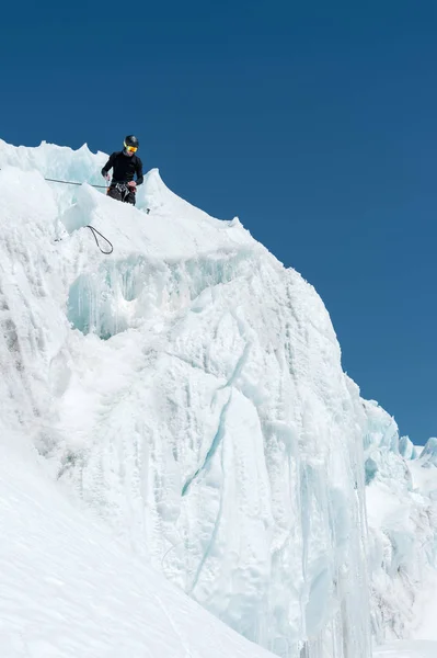 Um montanhista profissional em um capacete e máscara de esqui no seguro não entalhe o machado de gelo na geleira. O trabalho de um alpinista profissional no inverno em uma geleira contra o céu azul — Fotografia de Stock