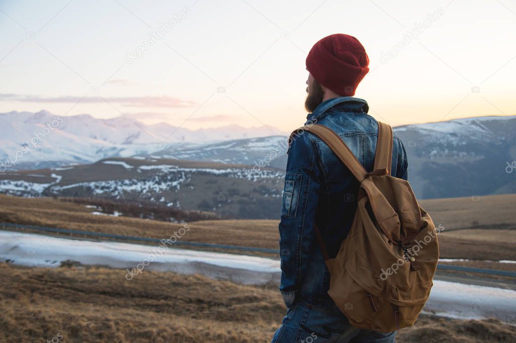 Bearded tourist hipster man in a hat with a backpack stand back on a roadside bump and watching the sunset against the background of a snow capped mountain