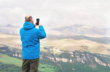 Sakallı bir adam bir membran mavi ceketli ve dağların fon karşı doğada standları telefon manzara fotoğraf çeker
