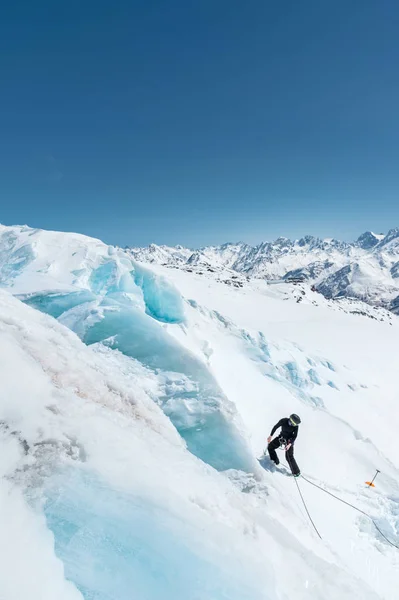 一个专业的登山运动员在头盔和滑雪面具保险在冰川上的一个缺口洞在高加索雪山的背景下 — 图库照片