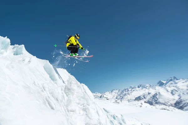 Vor dem Hintergrund des blauen Himmels und der kaukasischen schneebedeckten Berge springt ein Skifahrer in voller Sportausrüstung vom Gipfel des Gletschers in den Abgrund. elbrus region. Russland — Stockfoto