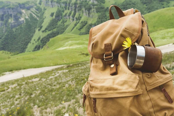 Mochila vintage amarilla Hipster con una taza fijada en ella con una taza vista frontal de cerca. Viajeros bolsa de viaje en el fondo de un paisaje de montaña — Foto de Stock