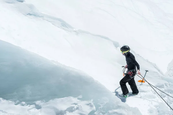 Un montañista profesional con casco y pasamontañas en el seguro hace un agujero en el glaciar contra el telón de fondo de las montañas caucásicas cubiertas de nieve — Foto de Stock