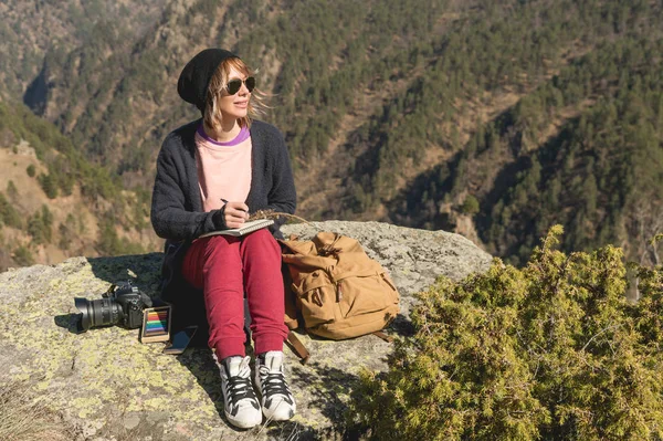 Стильна дівчина-дизайнер маляр в горах, що сидить на скелі малює в блокноті на заході сонця — стокове фото