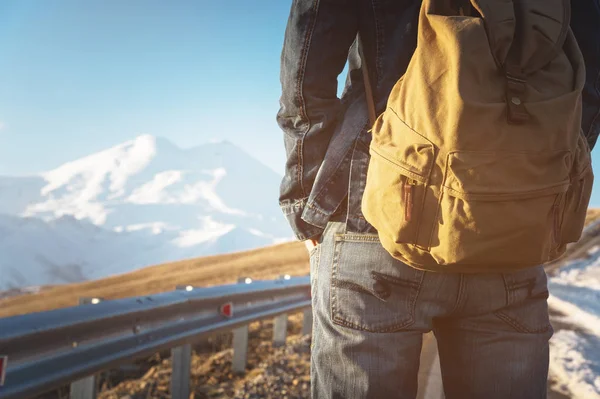 Primer plano de una mochila en la parte posterior de un viajero masculino caminando a lo largo de una carretera rural en el fondo de una montaña. Copia el espacio en blanco para el dieser. Viaje. — Foto de Stock