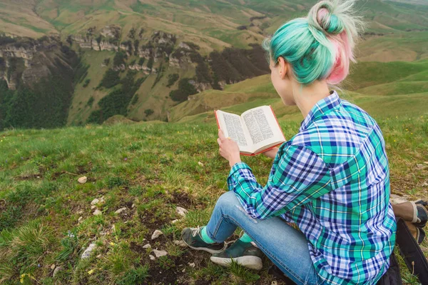 Ένα κορίτσι ταξιδιώτης κάθεται στα βουνά στο γρασίδι και διαβάζει ένα βιβλίο σχετικά με το ιστορικό έπος βουνά. Η έννοια της ανάγνωσης κατά τη διάρκεια της ανάπαυσης και διακοπές — Φωτογραφία Αρχείου
