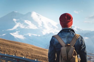 Sakallı turist hipster erkek güneş gözlüğü bir sırt çantası ile dimdik geri üzerinde yol kenarına yerleştirilen bir yumru ve şapkalı kar dağ çerçevede gün batımını izlemek