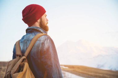 Sakallı turist hipster adam şapka ile bir sırt çantası içinde durmak geri yol kenarına yerleştirilen bir yumru ve şapkalı kar dağ çerçevede gün batımını izlemek