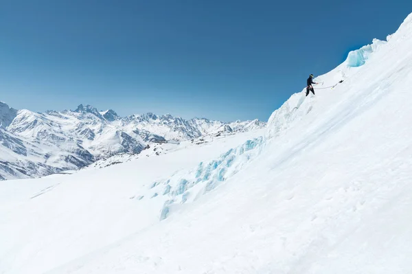 Um montanhista profissional em um capacete e máscara de esqui no seguro faz um buraco na geleira contra o pano de fundo das montanhas cobertas de neve caucasianas — Fotografia de Stock