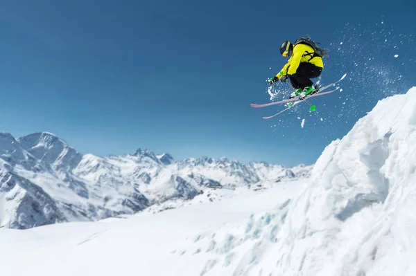 Um esquiador em equipamento desportivo completo salta para o precipício do topo da geleira contra o fundo do céu azul e as montanhas cobertas de neve caucasianas. Região de Elbrus. Rússia — Fotografia de Stock