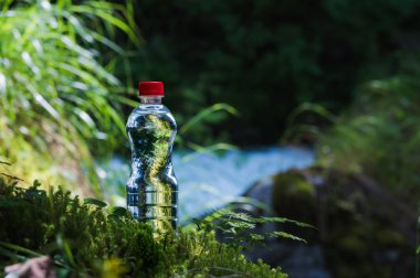 Şeffaf plastik A şişe kırmızı kapaklı temiz su standları çim ve moss bir engebeli dağ nehir arka plan üzerinde. Saf doğal içme suyu kavramı