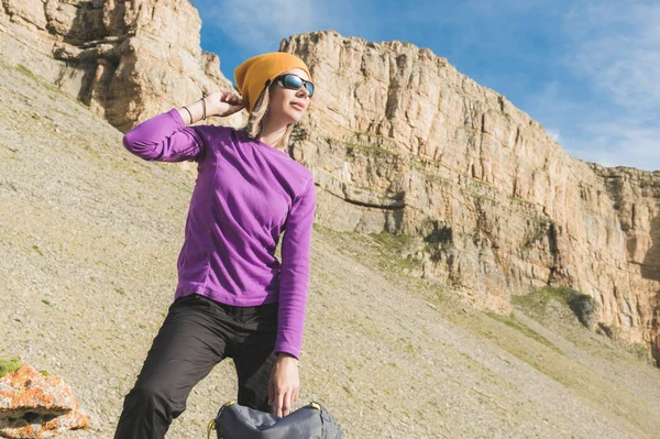 Усміхнена дівчина мандрівник в жовтому капелюсі і пара сонцезахисних окулярів стоїть біля підніжжя епічних скель з рюкзаком поруч і дивиться — стокове фото