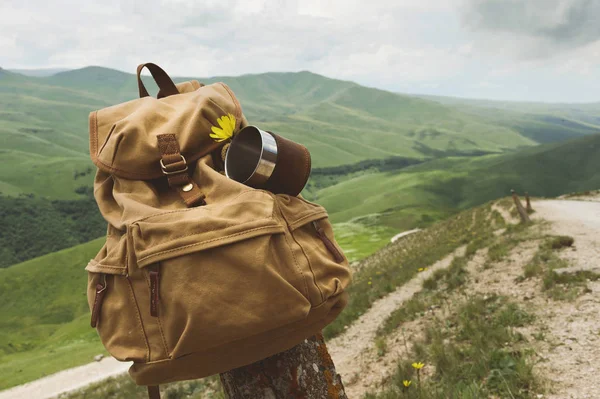 Mochila vintage amarilla Hipster con una taza fijada en ella con una taza vista frontal de cerca. Viajeros bolsa de viaje en el fondo de un paisaje de montaña y un camino — Foto de Stock