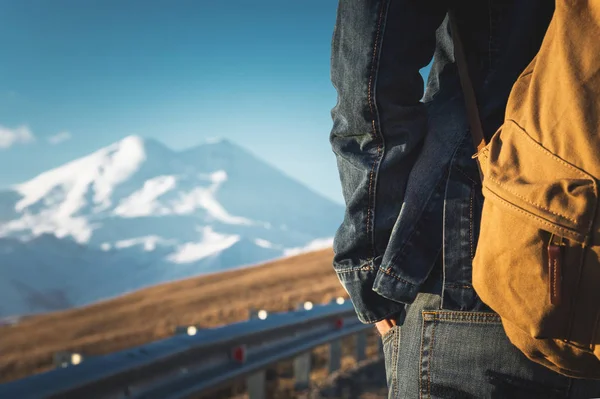 Primo piano di uno zaino sul retro di un viaggiatore di sesso maschile che cammina lungo una strada di campagna sullo sfondo di una montagna. Copia lo spazio vuoto per il moribondo. Viaggio — Foto Stock