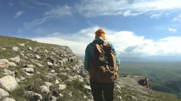 Девушка, которая ходит в шляпе сзади. Турист с рюкзаком прогуливается вдоль скалистого плато к утесу, чтобы полюбоваться видом. Slow Motion 4k — стоковое видео