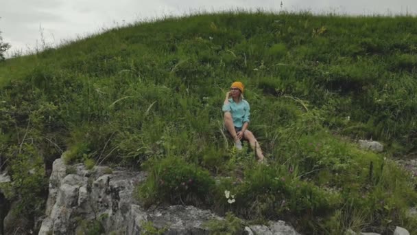 Jonge hipster vrouw zit op de rand van het gras van hoge muur van rots. Luchtfoto. Drone vliegt langzaam achteruit uit model. Tot oprichting van onthullende schot — Stockvideo