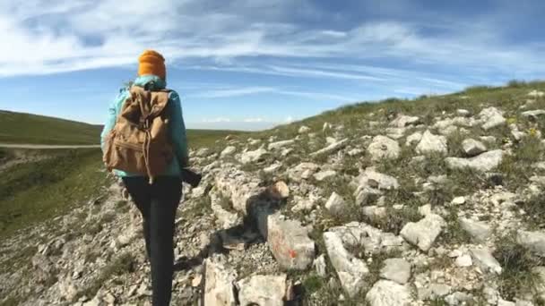 一个女孩从后面戴着帽子去拍照。一个带着背包和照相机的旅游摄影师正沿着一个岩石的高原走到悬崖上欣赏风景。慢 — 图库视频影像