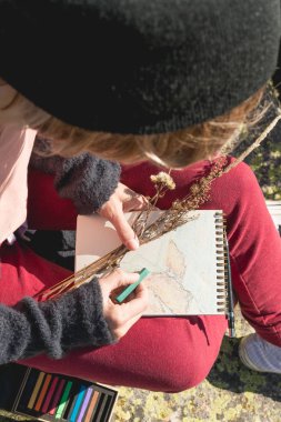 Şık kız tasarımcı ressam dağlarda bir kayaya oturan bir Not Defteri'nde günbatımında çizer.