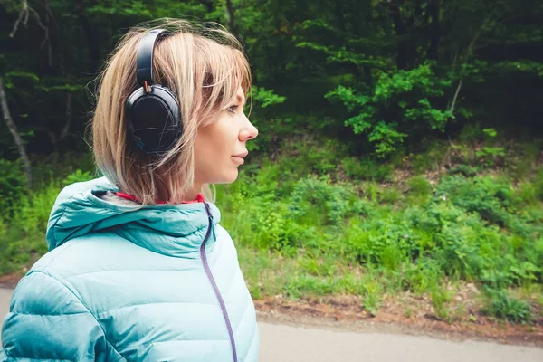 Portret Sporty atrakcyjny dziewczynka Blondynka w świetle działa w dół kurtka opatrunku bluetooth słuchawki z muzyką lub dźwięki natury na leśnej drodze — Zdjęcie stockowe