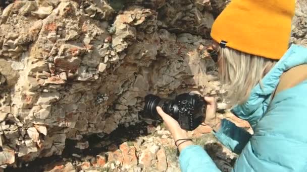 Una fotógrafa rubia con sombrero toma una foto en una pose extraña en su cámara digital con un fondo de rocas en el Cáucaso — Vídeo de stock