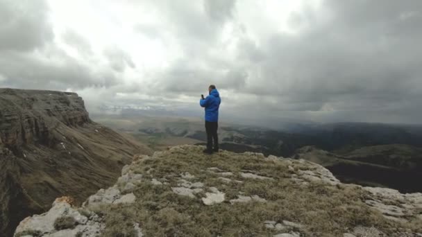 Hombre hipster barbudo toma fotos en su teléfono inteligente, mientras que en las rocas en las montañas del Cáucaso — Vídeos de Stock