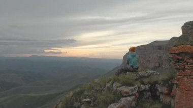 Hava - uçuş yetişkin kadın bir sırt çantası ile oturup geçmiş geriye dönük oturur ve rock epik Yaylası'nın eteklerinde günbatımında aittir. Arkadan görüntüleyin. Rusya. Kuzey Kafkasya