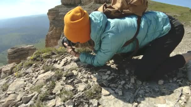 Блондинка фотограф в шляпе делает фотографию в странной позе на своей цифровой камере на фоне камней на Кавказе — стоковое видео