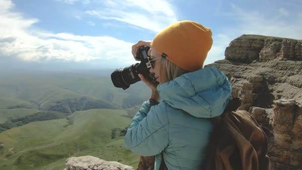 Chica rubia fotógrafa en la gorra toma una foto en su cámara digital con un fondo de rocas en el Cáucaso — Vídeo de stock
