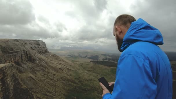 Hombre hipster barbudo toma fotos en su teléfono inteligente, mientras que en las rocas en las montañas del Cáucaso — Vídeos de Stock