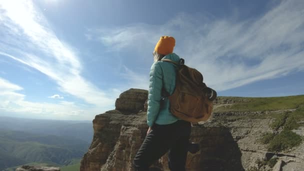 Una viaggiatrice sorridente con uno zaino e occhiali da sole si trova con la macchina fotografica sul bordo di un altopiano roccioso nel Caucaso — Video Stock