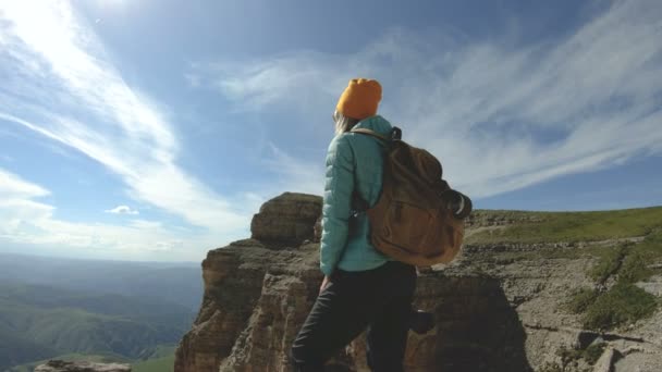 Uma menina viajante sorridente com uma mochila e usando óculos de sol fica com sua câmera na borda de um planalto rochoso no Cáucaso — Vídeo de Stock