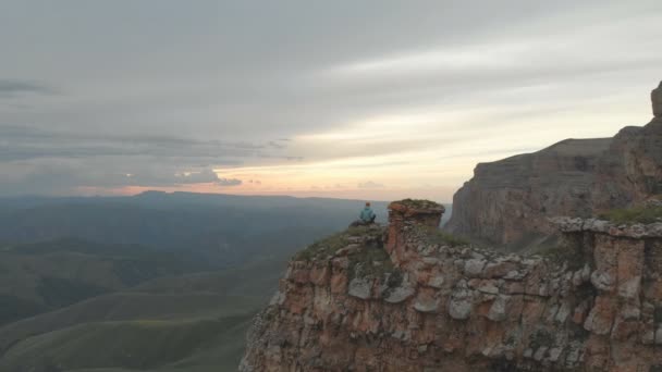 Aéreo - voo para a frente passado sentado em férias mulher adulta com uma mochila senta-se e descansa da rocha no sopé do planalto épico ao pôr do sol. Vista pelas traseiras. A Rússia. Norte do Cáucaso — Vídeo de Stock