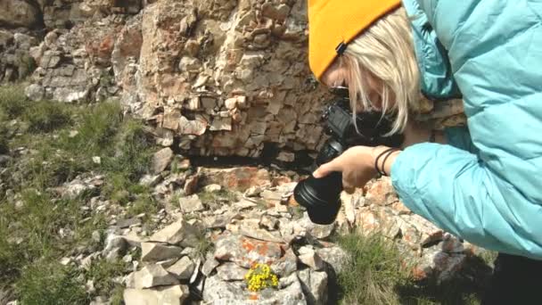 帽子の金髪の女の子の写真は コーカサス山脈の岩の背景と彼女のデジタル カメラに関する奇妙なポーズで写真を撮る — ストック動画