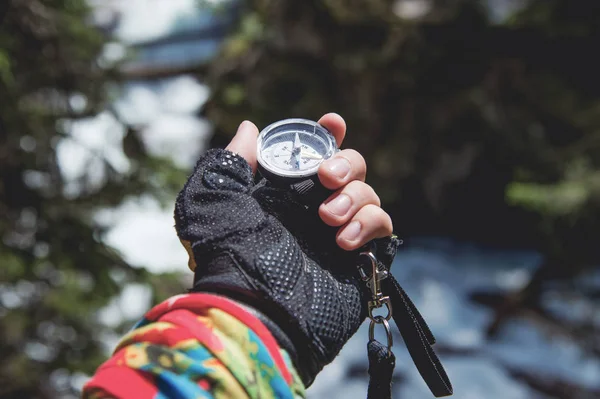 Een gehandschoende hand houdt een magnetisch kompas tegen de achtergrond van een naaldbos bos en berg rivier. Het concept van outdoor navigatie — Stockfoto