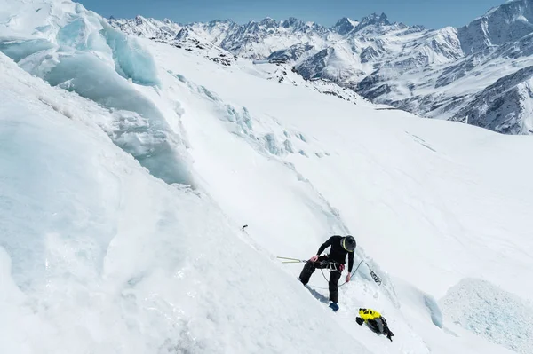 Профессиональный альпинист в шлеме и лыжной маске на страховке высекает ледяной топор в леднике. Работа профессионального альпиниста зимой на леднике против голубого неба — стоковое фото