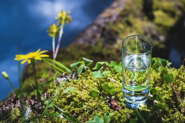 Clear water in a clear glass against a background of green moss with a mountain river in the background. Healthy food and environmentally friendly natural water