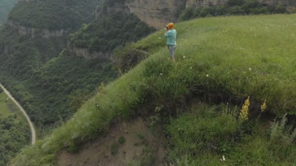 La fotógrafa en gafas y un sombrero toman una foto en su cámara dslr en el borde de la meseta cerca del precipicio. Vista aérea — Vídeos de Stock