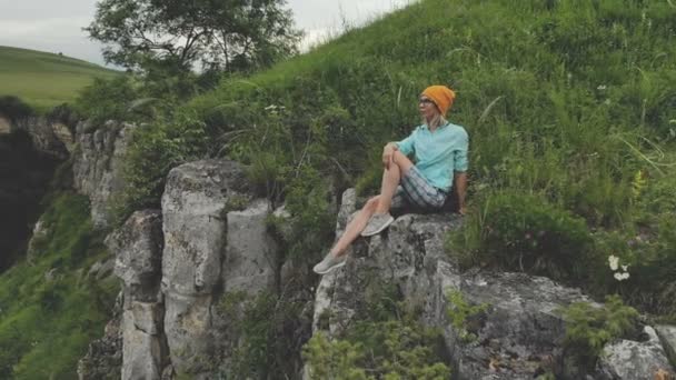 Young hipster woman is sitting on the edge on the grass of tall wall of rock. Aerial view. Drone is flying slow backward from model. Establishing revealing shot — Stock Video